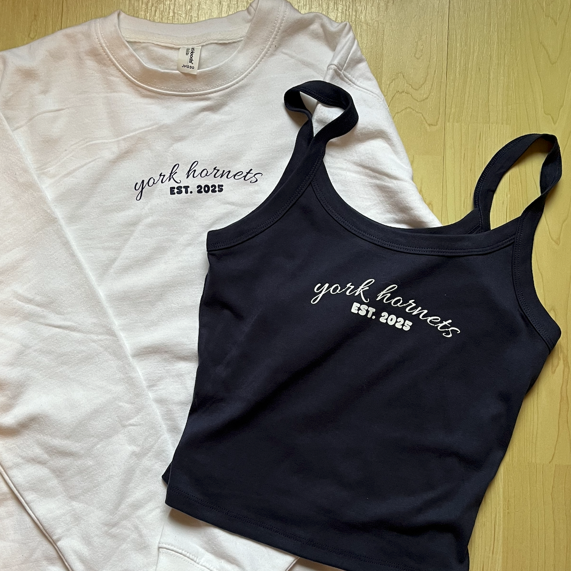A white crewneck sweatshirt and a black tank top, both featuring 'york hornets EST. 2025' text on the chest, laid flat on a light wood floor.