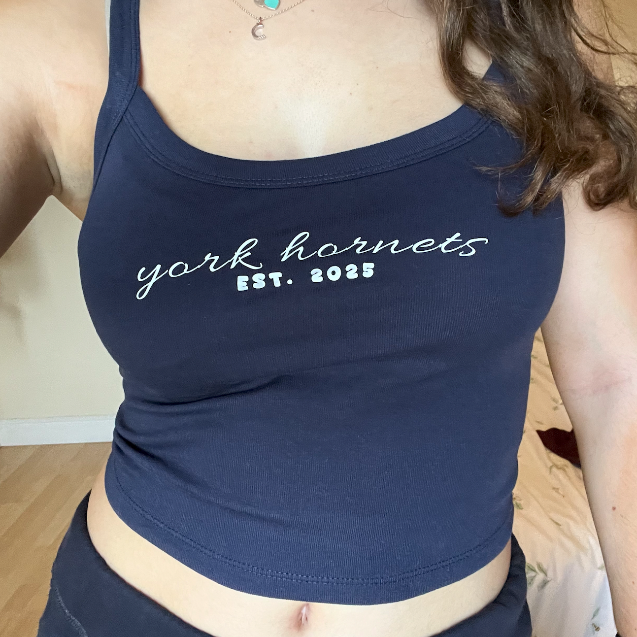 A woman wearing a navy blue scoop-neck crop top with 'york hornets EST. 2025' printed in white across the chest, shown in a selfie-style indoor photo.